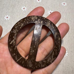 Könnte beinhalten: Ein rundes Friedenszeichen aus dunkelbrauner Kokosnussschale. Das Friedenssymbol ist aus der Mitte des Kreises ausgeschnitten. Das Naturmaterial hat eine strukturierte Oberfläche mit hellbraunen und weißen Flecken. Durchmesser ca. 8 cm.