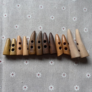 May include: A collection of wooden toggle buttons in various shades of brown, from light tan to dark walnut. Each button has a conical shape with two or three holes for sewing. The buttons are arranged on a gray fabric with a white and red floral pattern.