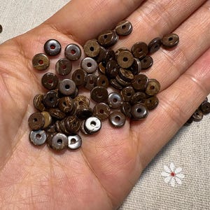 May include: A collection of small, round, brown beads with a hole in the centre. The beads are made of a natural material, possibly wood or coconut shell. The beads are scattered in a person's hand.