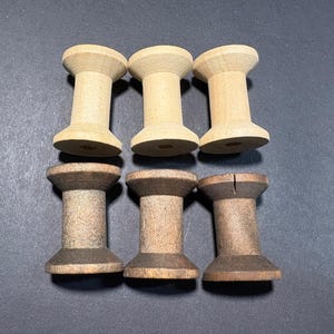 May include: Six wooden spools in two rows. The top row has three spools in a light wood colour. The bottom row has three spools in a darker wood colour. The spools are cylindrical with a wider top and bottom.