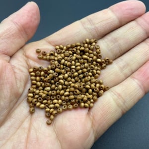 Puede incluir: Un puñado de pequeñas cuentas redondas de madera de color marrón dorado. Las cuentas están en la palma de una mano, con un fondo oscuro. Las cuentas parecen ser de tamaño y forma uniformes, adecuadas para la fabricación de joyas o manualidades.