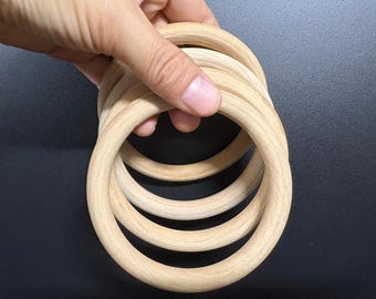 May include: A stack of unfinished wooden rings, held by a hand. The rings are light-colored, with a smooth, rounded shape. The background is a solid black.