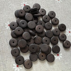 Könnte beinhalten: Eine Ansammlung dunkelbrauner, runder Holzperlen mit einem Loch in der Mitte. Die Perlen liegen auf einem hellen Stoff mit floralem Muster. Die Perlen sind unbehandelt und weisen eine natürliche Holzmaserung auf.