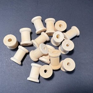 Puede incluir: Una colección de pequeñas bobinas de madera sin terminar. Cada bobina tiene un cuerpo cilíndrico con un agujero central y extremos ensanchados. Las bobinas son de color madera clara y están dispersas sobre una superficie oscura.