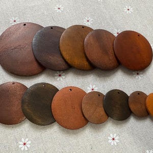 May include: A collection of wooden circles in various sizes and shades of brown, from dark to light. Each circle has a small hole at the top, suggesting they are designed for hanging or crafting. The arrangement showcases the natural wood grain.