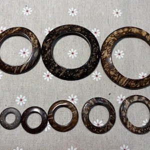 Puede incluir: Colecci&oacute;n de siete anillos de madera marr&oacute;n oscuro de diferentes tama&ntilde;os. Los anillos circulares tienen un aspecto natural y texturizado, con patrones de vetas de madera visibles. Est&aacute;n dispuestos sobre una tela gris claro con estampado floral.