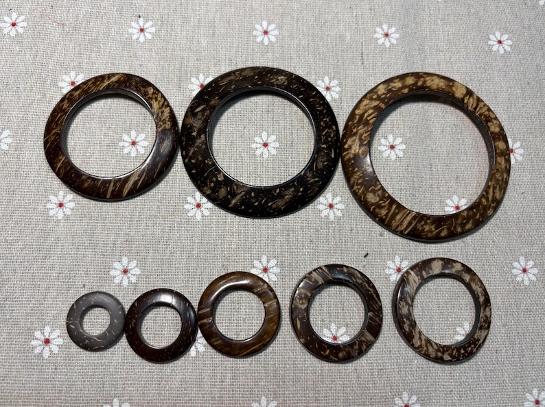 Puede incluir: Colecci&oacute;n de siete anillos de madera marr&oacute;n oscuro de diferentes tama&ntilde;os. Los anillos circulares tienen un aspecto natural y texturizado, con patrones de vetas de madera visibles. Est&aacute;n dispuestos sobre una tela gris claro con estampado floral.