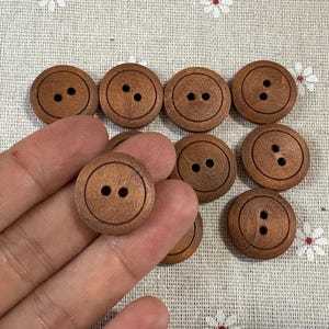 以下が含まれることがあります： さまざまなサイズの丸い木製ボタンのコレクション。各ボタンは温かみのある茶色で、縫い付け用の穴が2つまたは4つあります。ボタンは花柄の生地に表示されています。