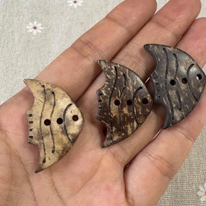 May include: Three fish-shaped buttons made from natural-colored coconut shell. The buttons have two holes for sewing and feature etched details to resemble fish scales and fins. The buttons vary in color from light tan to dark brown.