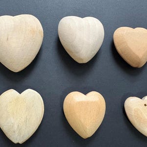 以下が含まれることがあります： 暗い表面に配置された、さまざまな明るい茶色とベージュの色合いの6つの木製のハート型のオブジェクト。 ハートは滑らかで彫刻されたテクスチャで、1つは穴が開いています。 これらの装飾的な木製のハートは、工芸品に適しています。