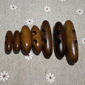 May include: A collection of seven wooden buttons in varying shades of brown. Each button is oval-shaped with two or three circular holes. The buttons are arranged on a floral patterned fabric.