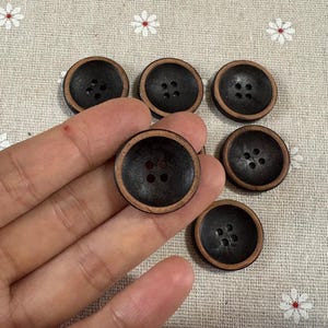 Puede incluir: Seis botones de madera marrón oscuro con un borde marrón claro, cada uno con cuatro agujeros. Los botones están dispuestos sobre una tela beige con estampados de flores blancas. Una mano sostiene algunos botones.