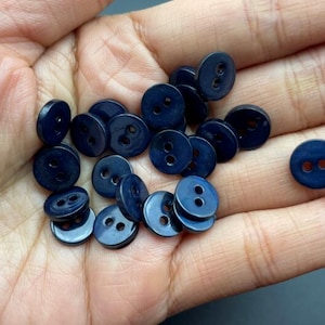 May include: A collection of navy blue, two-hole buttons. The buttons are round and have a slightly glossy finish. They are scattered across a hand, with some buttons stacked on top of each other.