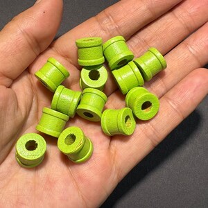 May include: A handful of small, lime green wooden beads. Each cylindrical bead has a central hole and a rounded shape. The beads are held in a hand, showing their size and potential use in crafts. The beads are approximately 1 cm in diameter.