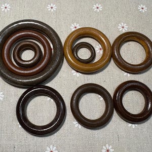May include: A selection of wooden rings in different sizes and shades of brown. The rings vary from dark to light brown, some with a glossy finish. The arrangement includes nested rings and individual rings, displaying the natural wood grain.