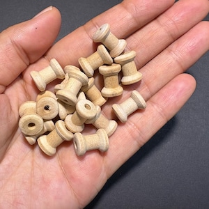 Puede incluir: Un conjunto de pequeñas bobinas de madera sin terminar, cada una con un cuerpo cilíndrico y un agujero central. Las bobinas se sostienen en una mano, mostrando su veta natural de madera y color marrón claro. Ideal para manualidades y proyectos de bricolaje.