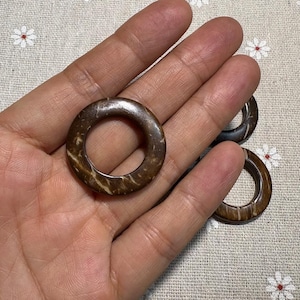 Puede incluir: Primer plano de varios anillos de coco marrones con un agujero en el centro. Los anillos tienen un aspecto natural y texturizado con tonos marr&oacute;n claro y oscuro. Los anillos se muestran sobre una tela con estampado floral.