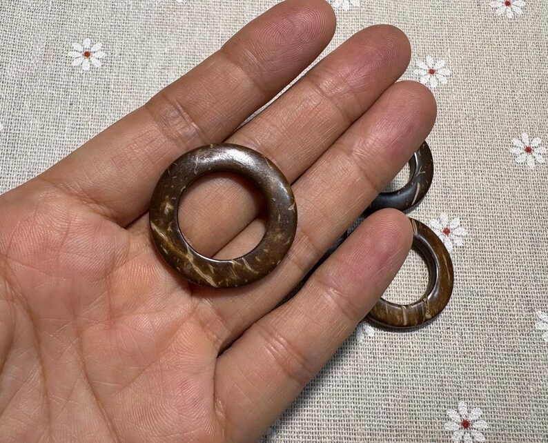 Puede incluir: Primer plano de varios anillos de coco marrones con un agujero en el centro. Los anillos tienen un aspecto natural y texturizado con tonos marr&oacute;n claro y oscuro. Los anillos se muestran sobre una tela con estampado floral.