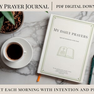 Może przedstawiać: Biały dziennik zatytułowany "My Daily Prayers" z zieloną obwódką, obok filiżanki kawy, długopisu i rośliny na marmurowej powierzchni. Tekst "Start each morning with intention and peace" znajduje się na dole obrazu.