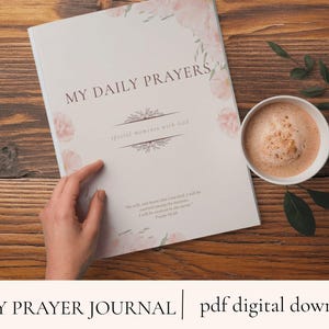 Może przedstawiać: Dziennik zatytułowany "My Daily Prayers" z kwiatowymi akcentami i tekstem "special moments with God". Obok dziennika na drewnianej powierzchni stoi filiżanka kawy. Dziennik jest otwarty, a ręka wskazuje stronę.