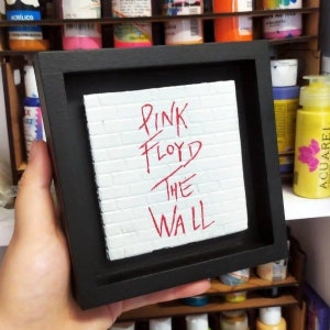 Puede incluir: Una pieza de arte enmarcada con un fondo de ladrillo blanco y las palabras "Pink Floyd The Wall" escritas en rojo.