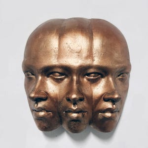 Puede incluir: Escultura de color bronce que representa tres rostros fusionados. Los rostros están de perfil, con rasgos detallados como ojos, narices y bocas. La escultura tiene un acabado liso y metálico, sobre un fondo blanco.