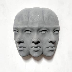以下が含まれることがあります： グレーのコンクリート彫刻で、3つの顔が並んでいます。各顔は目を閉じ、特徴がはっきりしています。彫刻は白い壁に取り付けられており、ミニマリストな美学を作り出しています。