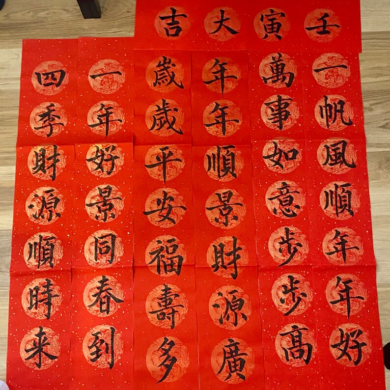 May include: Twenty red paper scrolls with black Chinese calligraphy. The scrolls are arranged in four rows of five. Each scroll has a different character written on it. The characters are: "吉", "大", "寅", "萬", "事", "如意", "步步", "高", "年", "年", "順", "景", "財源", "廣", "歲", "歲", "平安", "福壽", "多", "一年", "好景", "同", "春", "到", "四季", "財源", "順", "時", "來".