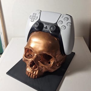 Puede incluir: Una escultura de calavera de color cobre con un mando de videojuegos blanco y negro encima. La calavera se asienta sobre una base cuadrada negra, creando una pieza decorativa única.
