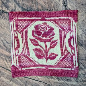 Könnte beinhalten: Ein quadratisches Strickstoffmuster mit einem zweifarbigen Farbschema in Magenta und Creme. Das Design zeigt ein zentrales Rosenmotiv in einem geometrischen Rahmen, umgeben von einem strukturierten, gesprenkelten Muster auf der gesamten Fläche.