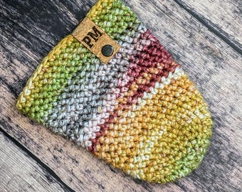 May include: A crocheted cup cozy with a multi-colored striped pattern in shades of green, yellow, white, and red. The cozy has a brown leather tag with the initials "PM" on it.