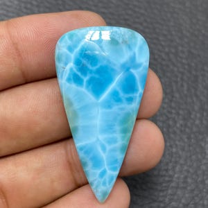 Può includere: Una gemma di Larimar lucidata a forma di goccia, caratterizzata da un vibrante colore azzurro cielo con intricati motivi bianchi a ragnatela sulla sua superficie liscia e lucida. La pietra presenta una base appuntita e un bordo superiore arrotondato.