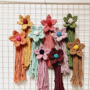May include: A collection of handmade macrame flower wall hangings in various colors, including orange, pink, green, and brown. Each flower has a central knot and cascading fringe, creating a textured, decorative display.