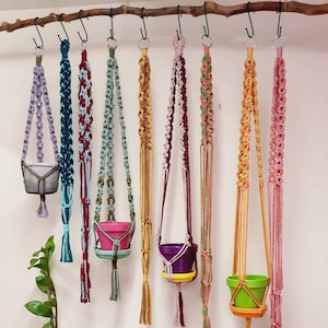 May include: Eight colorful macrame plant hangers with different colored pots hanging from a wooden branch. The hangers are made of cotton cord and have a variety of patterns. The pots are made of plastic and are in various colors, including purple, green, and orange.