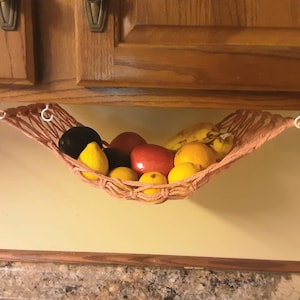 May include: A pink hammock-style fruit basket hanging from the underside of a wooden cabinet. The basket is filled with lemons, limes, oranges, bananas, and avocados.