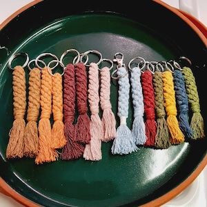 May include: A collection of 15 colorful keychains with tassels. The keychains are made of braided rope and come in a variety of colors, including brown, orange, red, pink, blue, green, and yellow.