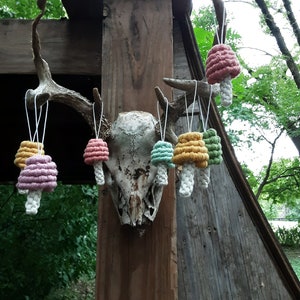 May include: A collection of seven crocheted mushroom ornaments in various colors, including pink, green, yellow, and orange, hanging from a deer antler on a wooden surface.