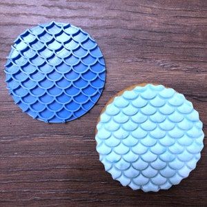 May include: A blue, circular cookie stamp featuring a mermaid scale design, alongside a cookie decorated with light blue icing in the same pattern. The stamp is plastic, and both items rest on a wooden surface. The cookie has a light brown edge.