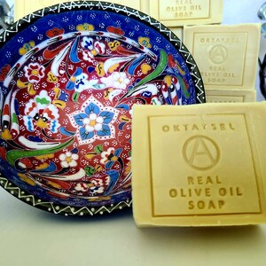 May include: A bar of natural olive oil soap with the words "OKTAYSEL" and "REAL OLIVE OIL SOAP" printed on the front. The soap is sitting on a white surface next to a decorative bowl with a colorful floral pattern.