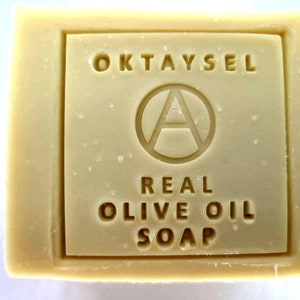 May include: A bar of Oktaysel Real Olive Oil Soap. The soap is white and has a rectangular shape. The brand name is printed on the front of the soap in a black font.