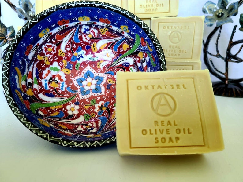 May include: A bar of natural olive oil soap with the words "OKTAYSEL" and "REAL OLIVE OIL SOAP" printed on the front. The soap is sitting on a white surface next to a decorative bowl with a colorful floral pattern.