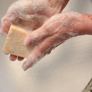 May include: A person's hands are covered in white soap suds as they rub a rectangular bar of soap.