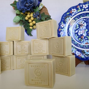 May include: A stack of rectangular bars of natural olive oil soap, labeled "OKTAYSEL REAL OLIVE OIL SOAP", on a white surface. The soap is arranged in a pyramid shape with the top bar centered. The soap is a light beige color and has a slightly textured surface.