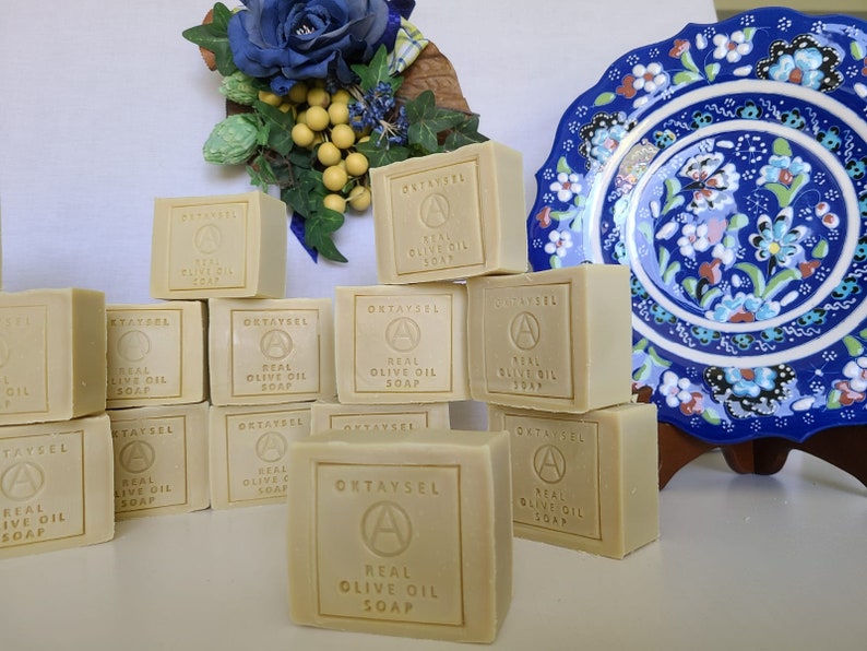 May include: A stack of rectangular bars of natural olive oil soap, labeled "OKTAYSEL REAL OLIVE OIL SOAP", on a white surface. The soap is arranged in a pyramid shape with the top bar centered. The soap is a light beige color and has a slightly textured surface.