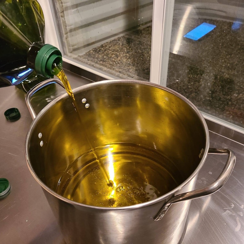 May include: A stainless steel pot with a handle being filled with golden yellow olive oil from a green glass bottle.