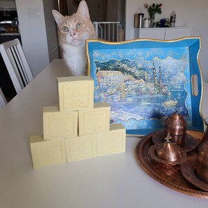 May include: A stack of eight rectangular bars of ivory-colored soap on a white table. The soap bars have gold lettering on them. A blue tray with a gold trim and a painted scene of a city on the water is in the background. A copper tray with a set of Turkish coffee cups is also in the background.