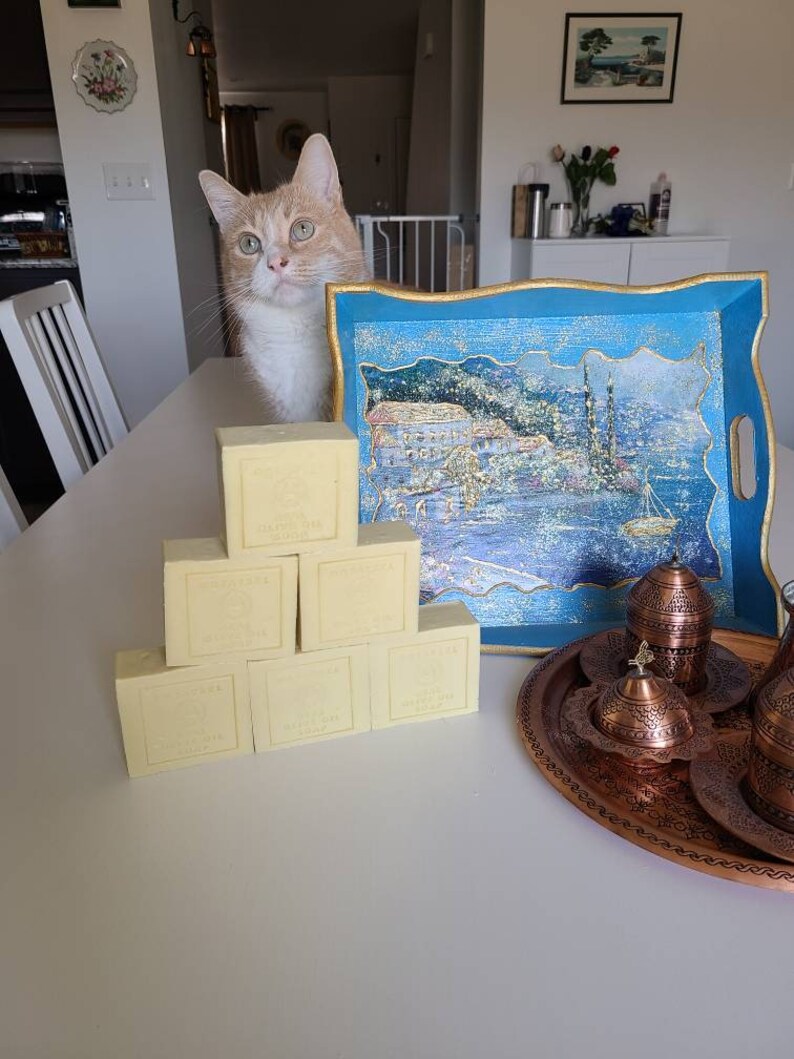 May include: A stack of eight rectangular bars of ivory-colored soap on a white table. The soap bars have gold lettering on them. A blue tray with a gold trim and a painted scene of a city on the water is in the background. A copper tray with a set of Turkish coffee cups is also in the background.