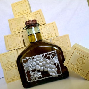 May include: A stack of yellow bars of soap with the text "OKTAYSEL REAL OLIVE OIL SOAP" on each bar. A glass bottle of olive oil with a silver grapevine design is in front of the soap.