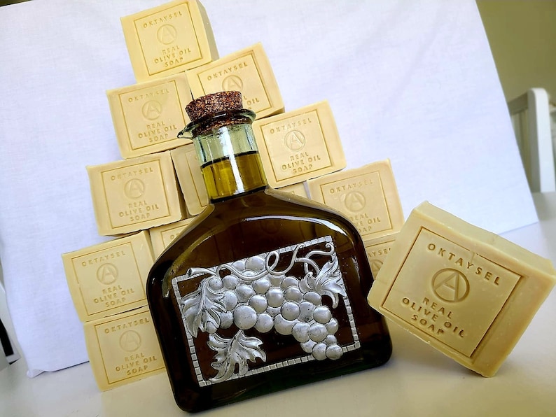 May include: A stack of yellow bars of soap with the text "OKTAYSEL REAL OLIVE OIL SOAP" on each bar. A glass bottle of olive oil with a silver grapevine design is in front of the soap.