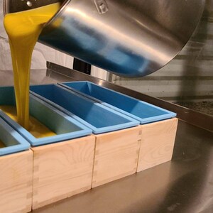 May include: A person pouring yellow liquid into five blue silicone molds, each sitting in a wooden box. The molds are lined up on a stainless steel surface.